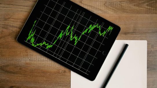 Flatlay of an iPad displaying stock market graph on a wooden desk with a pencil and paper.