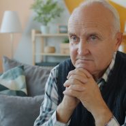Elderly man sitting on a couch with hands clasped.