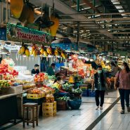 two people walking through a market with fruit and vegetables