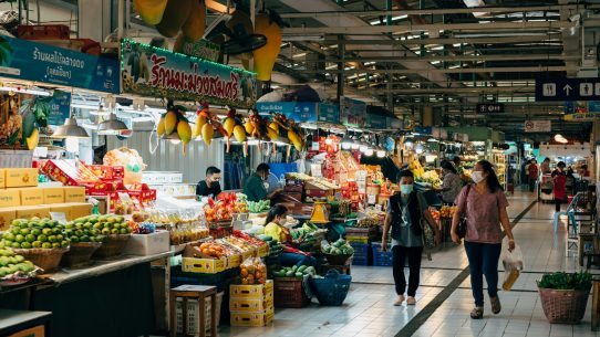 two people walking through a market with fruit and vegetables