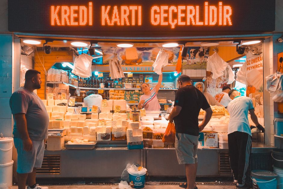 Turkish market stall with cheeses and customers under a bright credit card sign.
