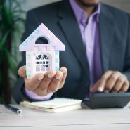 man holding model house at desk with calculator