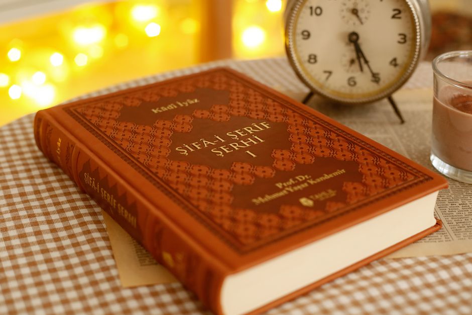 A vintage book next to an antique clock on a checkered tablecloth, with warm, ambient lighting.