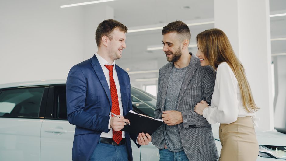 A smiling couple buys a new car from a confident salesman inside a modern car dealership.
