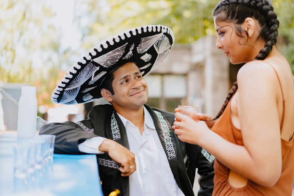 A couple dressed in traditional Mexican clothing smiling outdoors, enjoying a drink.